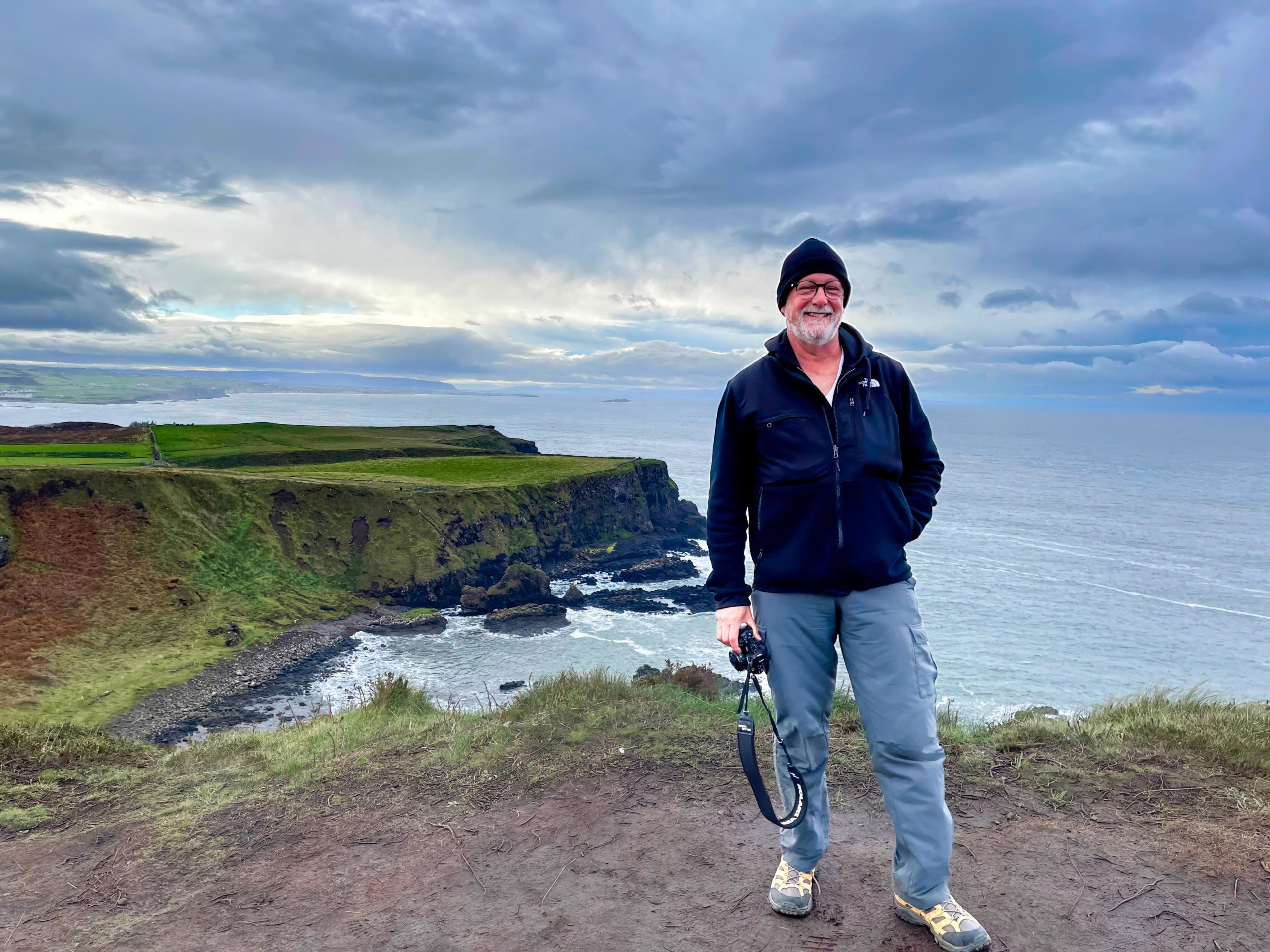 A photo of the author standing on top of a cliff in County Antrim, Northern Ireland. The sea is behind him and he is holding a camera in his hand.